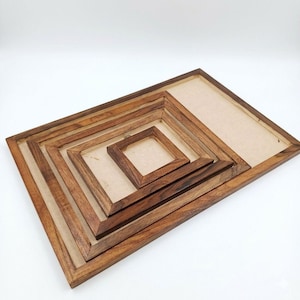Puede incluir: Un conjunto de cinco marcos de fotos de madera de diferentes tamaños, anidados entre sí. Los marcos son de color marrón cálido con vetas de madera natural. El marco más pequeño es cuadrado, el más grande es rectangular. El fondo es de color beige claro.