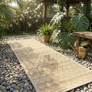 May include: A rectangular, light-colored bamboo runner rug with a diamond pattern, laid on a bed of pebbles. The rug leads towards a wooden gazebo, surrounded by lush greenery, including large-leafed plants and potted ferns. A brass watering can sits nearby.