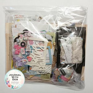 May include: A clear plastic bag containing a collection of craft supplies, including paper ephemera, die-cut shapes, pink and purple bows, a white frame cutout with the text "Hey Girls Hey", and a small pouch of white lace and assorted charms.