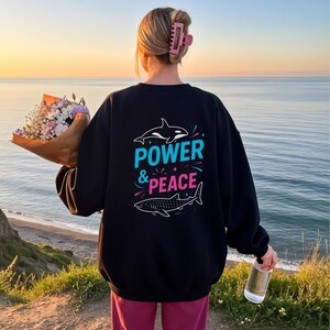 Può includere: Felpa nera con le parole "POWER & PEACE" in turchese e rosa, insieme a illustrazioni di un'orca e di uno squalo balena. La persona tiene in mano un mazzo di fiori e una bottiglia d'acqua, in piedi su una scogliera che si affaccia sull'oceano.