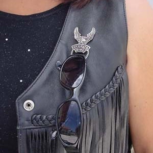 May include: A black leather vest with fringe detailing and a silver eagle pin with the text "Live to Ride". A pair of black sunglasses are clipped to the vest.