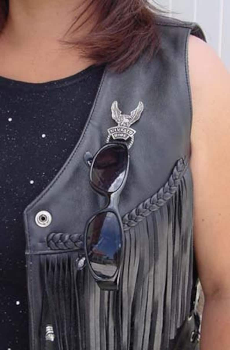 May include: A black leather vest with fringe detailing and a silver eagle pin with the text "Live to Ride". A pair of black sunglasses are clipped to the vest.