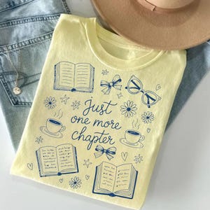 以下が含まれることがあります： クルーネックの明るい黄色のTシャツ。青いグラフィックデザインで、開いた本、コーヒーカップ、リボン、メガネ、花、星、そして「Just one more chapter」というテキストが描かれています。