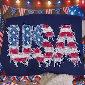 May include: A navy blue textile with the word "USA" appliqued in red, white, and blue fabric, resembling the American flag. The letters have a distressed, frayed edge design. The background includes festive decorations.