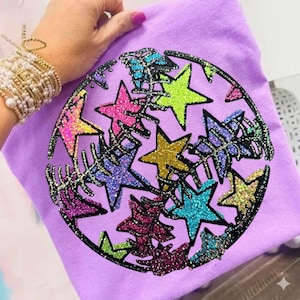 Pode incluir: Uma camiseta lilás clara com um design brilhante de estrelas em várias cores, incluindo rosa, amarelo, azul e verde, dispostas em um padrão circular. O design é delineado em preto.