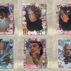 Puede incluir: Seis tarjeteros rectangulares de plástico decorados con varias pegatinas, pedrería y detalles holográficos. Cada soporte contiene una fotografía de retrato de una persona, con temas de colores rosa, azul y morado con adornos.