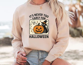 May include: Beige sweatshirt with a Halloween graphic. The graphic features a smiling jack-o'-lantern, a black cat wearing a witch hat, and a friendly ghost. The text reads "IT'S NEVER TOO EARLY FOR HALLOWEEN."