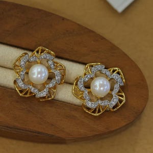 May include: A pair of stud earrings featuring a central white pearl surrounded by a gold-toned, open-work floral frame encrusted with small, clear, sparkling stones. The design creates a layered, petal-like appearance around the pearl.