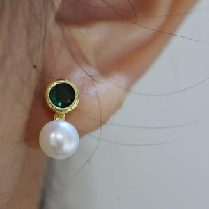 Puede incluir: Un pendiente de tono dorado que presenta una piedra preciosa redonda de color verde oscuro engastada, situada directamente sobre una perla esférica blanca y lisa. El pendiente tiene un diseño colgante con una montura metálica sencilla.