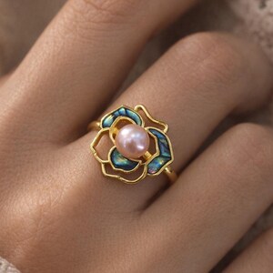 May include: A gold ring with a floral design. The ring features a pink pearl at the centre, framed by blue and green iridescent elements. The ring is on a finger.