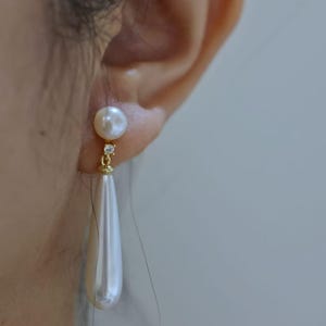 Könnte beinhalten: Nahaufnahme eines goldfarbenen Ohrrings mit einem weißen Perlenstecker und einer tropfenförmigen Perle zum Anhängen. Der Ohrring hat einen kleinen, klaren Edelstein-Akzent. Der Hintergrund ist eine verschwommene, neutrale Farbe.