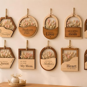 Könnte beinhalten: Eine Sammlung von Holz-Wandbehängen mit Trockenblumen und verschiedenen eingravierten Botschaften. Die Formen umfassen Sechsecke, Kreise und Bögen. Einige haben Texte wie "Danke" und "Muttertag". Die Rahmen sind hellbraun.