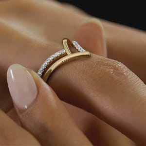 May include: A gold-colored ring with a unique design, featuring a curved band and a vertical bar. The ring is adorned with small, sparkling gemstones along the band and the top of the vertical bar, creating a dazzling effect. The ring is worn on a finger.