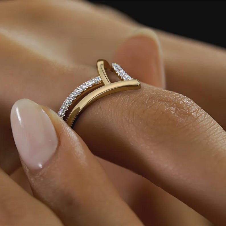 May include: A gold-colored ring with a unique design, featuring a curved band and a vertical bar. The ring is adorned with small, sparkling gemstones along the band and the top of the vertical bar, creating a dazzling effect. The ring is worn on a finger.