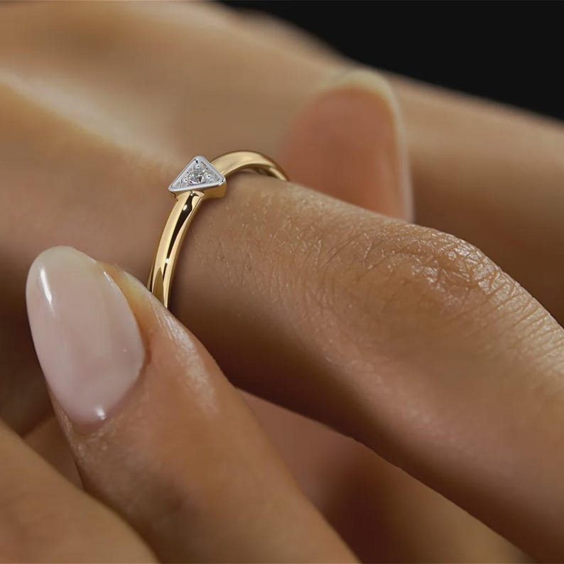 May include: A gold ring with a triangular, diamond-studded centerpiece. The ring is on a finger, showcasing its delicate design and the sparkle of the diamonds. The band is a polished gold, creating a contrast with the white diamonds.