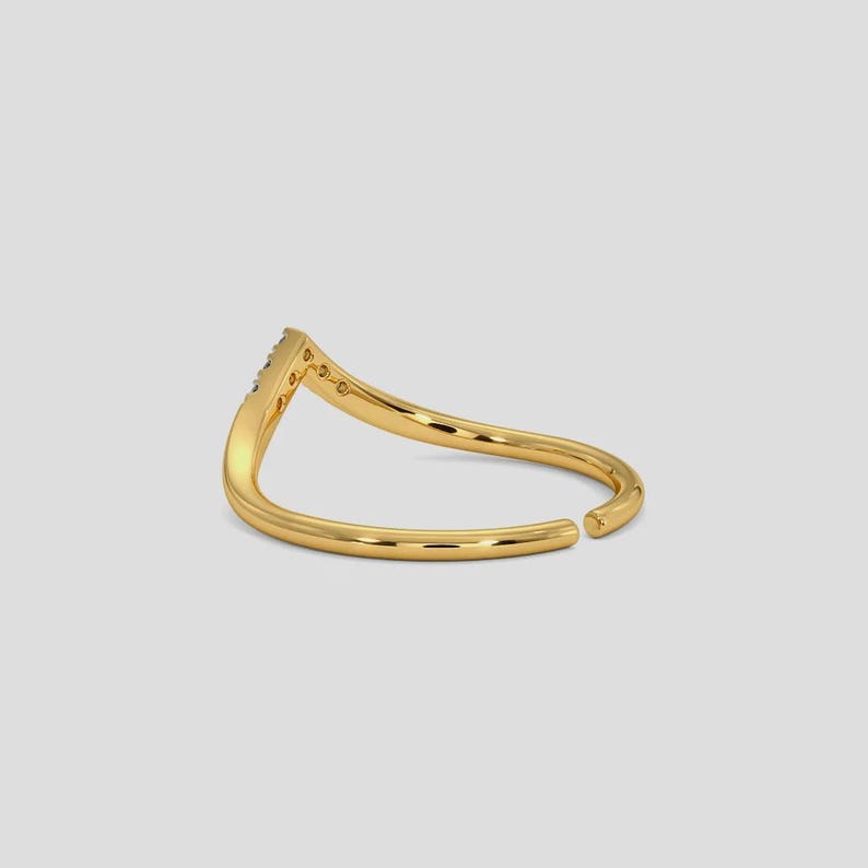 May include: A gold-colored ring with a V-shaped design. The ring features small, clear stones set along the top of the V. The band curves gracefully, with an open design at the bottom. The ring is set against a plain, light background.