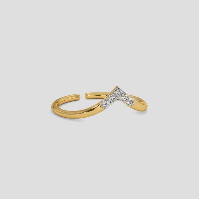 May include: A gold-colored ring with a V-shaped design. The ring features small, clear gemstones set along the V-shape. The ring is open at the bottom, allowing for size adjustment. The ring is set against a plain, light gray background.