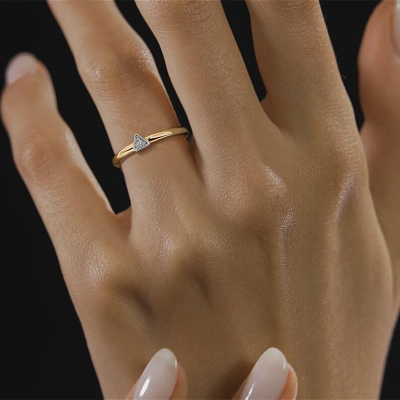 May include: A gold ring with a small, triangular, diamond-studded design. The ring is on a finger, showcasing the delicate band and the sparkling gemstone. The ring is a piece of jewelry.