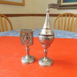 May include: Two silver-toned metal religious ceremonial items on a red table cloth. The item on the left is a square box with a decorative design. The item on the right is a tall, round container with a decorative design and a small flag on top.