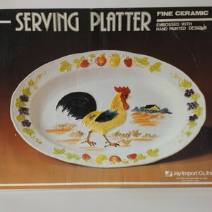 May include: A white oval serving platter with a hand-painted design of a rooster, fruit, and grapes. The platter is made of fine ceramic and is embossed with the words "Serving Platter" and "Fine Ceramic Embossed with Hand Painted Designs". The platter is packaged in a brown box with the company name "Jay Import Co., Inc."