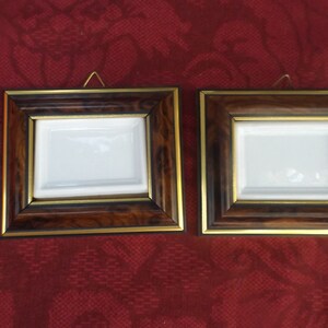 May include: Two square picture frames with a dark wood grain finish and gold trim. Each frame has a white interior and a small brass hanger. The frames are displayed against a red patterned background.