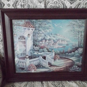 May include: A framed painting depicting a white building with a porch overlooking a blue body of water. The building has a white railing and a white door. There are trees and flowers in the foreground. The painting is in a dark brown frame.