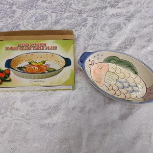 May include: A hand-painted underglaze table plate with a fish design. The plate is oval shaped and has two blue handles. The fish is painted in shades of blue, yellow, orange, and pink.