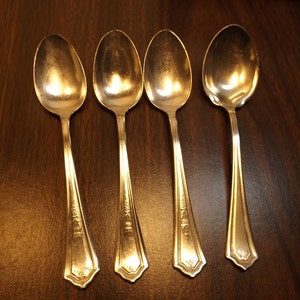 May include: Four silver spoons arranged in a row. The spoons have oval bowls and ornate, detailed handles. The spoons are made of a shiny, reflective metal, likely silver or a silver-colored alloy. The spoons are set against a dark wooden surface.