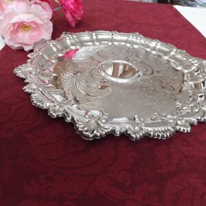 May include: A silver serving tray with an ornate, embossed design. The tray has a raised center and a scalloped edge.