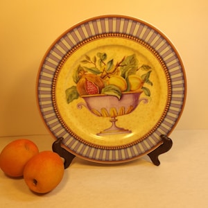 May include: A decorative ceramic plate with a blue and gold border. The center of the plate features a painted image of a bowl filled with fruit, including figs, pears, and lemons.