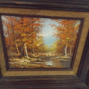 May include: A framed oil painting depicting a forest scene with trees in autumn colors. The trees are in the foreground with a small stream in the middle ground. The background is a hazy blue sky.