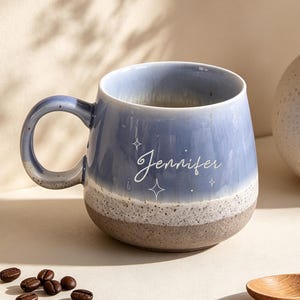 Può includere: Una tazza in ceramica con la parte superiore smaltata in blu e la base beige maculata. La tazza presenta il nome Jennifer in caratteri bianchi in corsivo, accentuato da piccole icone a forma di stella bianca. Ha un corpo rotondo e un manico.