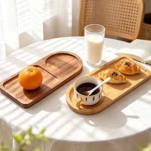 Puede incluir: Dos bandejas de servicio rectangulares de madera con extremos redondeados. Una bandeja contiene una naranja, mientras que la otra sostiene una taza de cerámica con una cuchara blanca y dos cruasanes. Un vaso alto de leche está entre ellas.