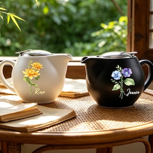 May include: A white teapot with a yellow flower illustration and the name Rose, and a black teapot with a blue and purple flower illustration and the name Jennie. Both teapots feature a rounded body, a side handle, and a metallic lid.