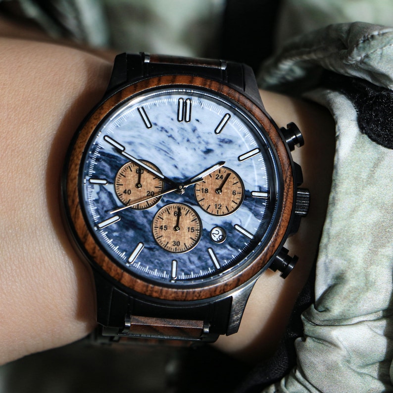 May include: A wristwatch with a black case and a wooden bezel. The watch face has a blue and white marble design with three smaller dials in a tan color. The watch is on a person's wrist.