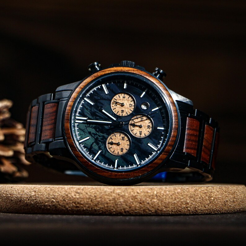May include: A black and wood-toned chronograph watch with a dark blue face and three sub-dials. The watch features a wooden bezel and band, and is displayed on a cork surface. The watch has a classic, elegant design.
