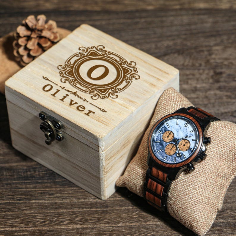 May include: A wooden watch with a brown and black band and a light blue face with orange sub-dials rests on a burlap pillow. A wooden box with the name "Oliver" and the letter "O" engraved on the lid sits next to the watch.