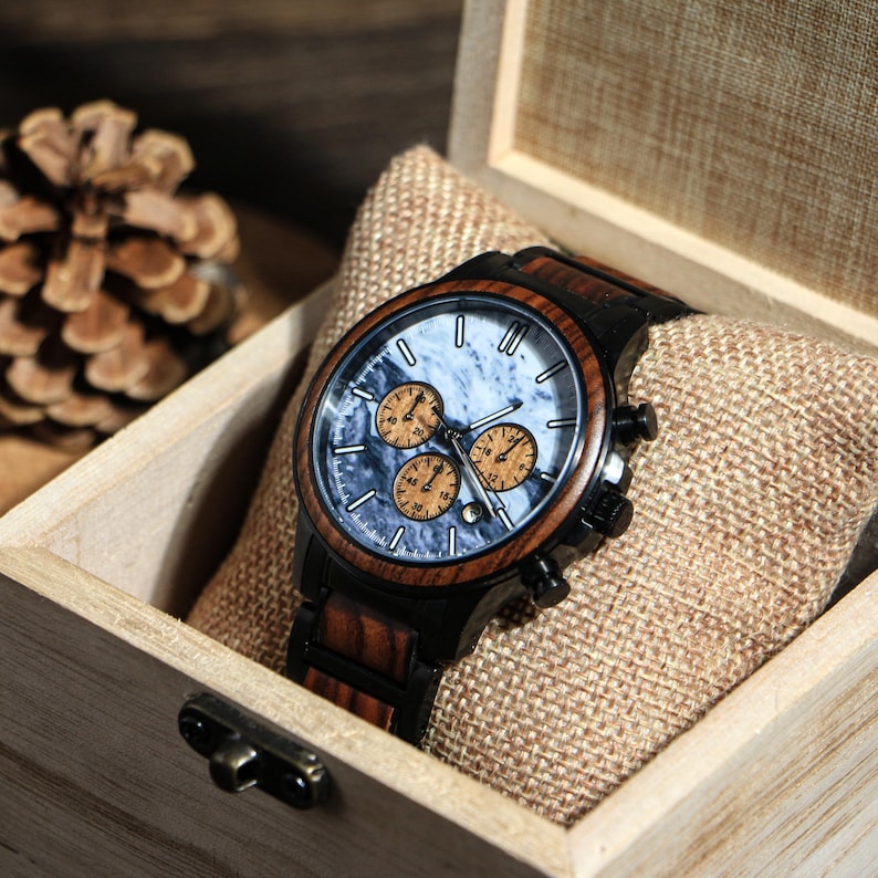 May include: A wooden wristwatch with a marble-patterned face and three sub-dials, set in a wooden box. The watch has a dark wood bezel and a black band. The watch is displayed on a tan fabric pillow.
