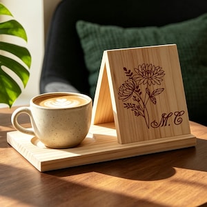 May include: A light-colored wooden tray with a circular indentation holding a speckled ceramic mug filled with latte art. An upright wooden panel attached to the tray features a dark red floral line drawing and the letters M.C. engraved on it.