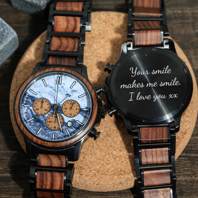 May include: Two wooden watches with black metal accents. One watch has a blue and white marbled face with three sub-dials. The other watch has the inscription "Your smile makes me smile. I love you xx" on the back.