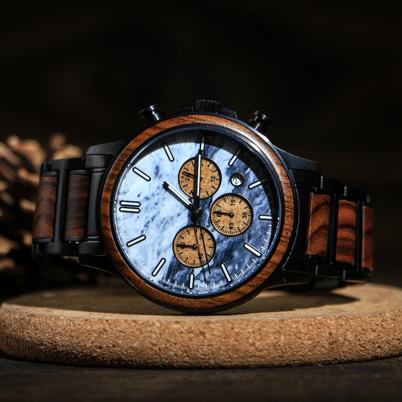 May include: A wooden wristwatch with a black case and a marble-patterned face. The watch features three sub-dials and a date window. The band is a combination of black metal and wood. The watch is displayed on a small, round wooden stand.