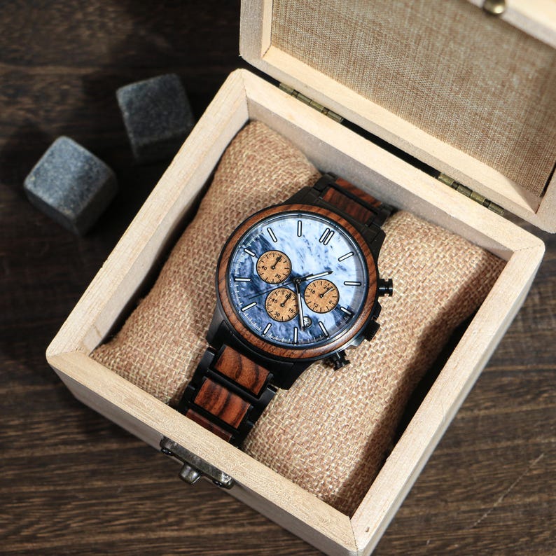 May include: A wooden wristwatch with a blue and orange face, presented in a wooden box with a burlap lining. The watch features a black bezel and a wooden band. Two gray stone cubes are visible.
