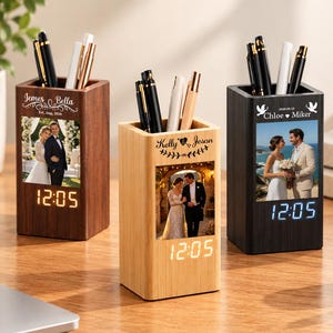 May include: Three rectangular wooden pen holders with integrated digital clocks. Each holder features a custom printed photograph of a couple, personalised names, and dates. The holders are filled with various pens and are finished in dark wood, light wood, and black wood tones.