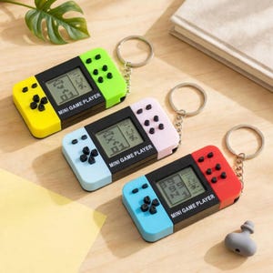 Puede incluir: Tres llaveros rectangulares de mini consola de juegos con pantallas negras y botones. Uno es amarillo y verde lima, otro azul claro y rosa pálido, y otro azul claro y rojo. Cada uno tiene una anilla metálica y el texto MINI GAME PLAYER.
