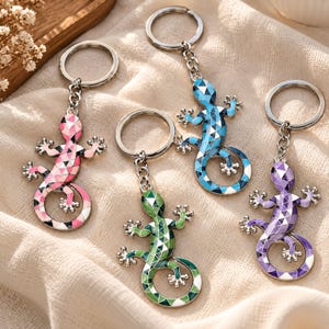 May include: Four metal keychains featuring lizard-shaped charms with a geometric mosaic pattern. Each lizard is filled with enamel in distinct colours: pink, blue, green, and purple. The charms are attached to silver-toned split rings by short chains.