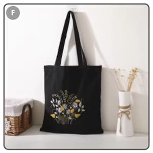 Puede incluir: Bolso tote negro con dise&ntilde;o floral bordado en amarillo, blanco y verde. El bolso tiene asas largas negras y est&aacute; hecho de una tela resistente. El bolso tote se muestra contra una pared blanca.