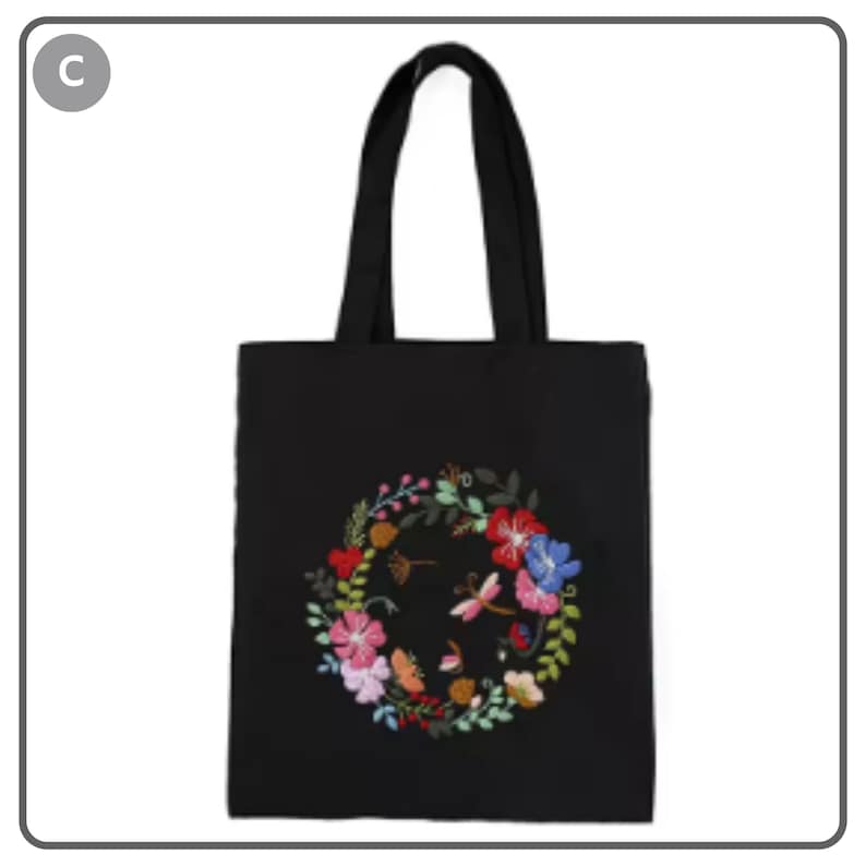 Puede incluir: Bolso tote negro con un dise&ntilde;o de bordado floral. El bordado presenta una corona de flores coloridas, incluyendo flores rojas, rosas y azules, con una lib&eacute;lula en el centro. El bolso tiene asas negras.