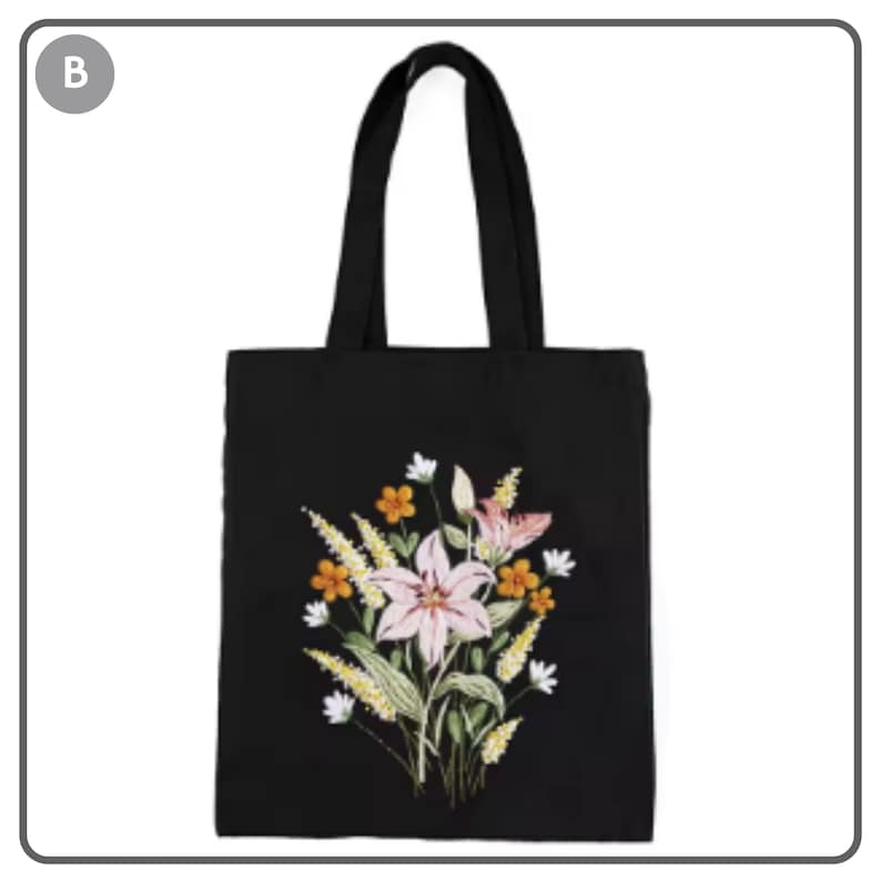 Puede incluir: Bolso tote negro con dise&ntilde;o floral bordado. El dise&ntilde;o presenta un gran lirio rosa, flores naranjas y blancas y hojas verdes. El bolso tiene dos asas negras.