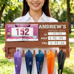 May include: Wooden medal display board with the name "Andrew's" and personal best times for 5K, Half, and Full marathons. Includes a race bib and hooks for hanging medals. The board is brown and holds several colorful ribbons.