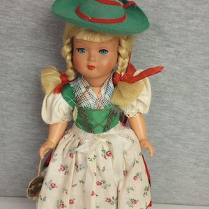 May include: A vintage doll wearing a traditional German outfit. The doll has blonde hair in braids, blue eyes, and red lips. She wears a green hat with a red band, a white blouse, and a floral skirt with red trim.
